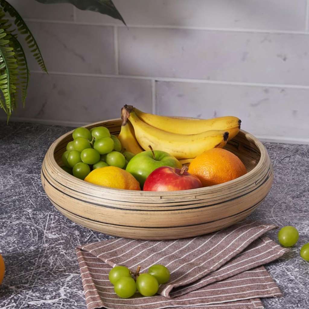 Natural bamboo serving bowl filled with fresh fruits including green grapes, bananas, apples, and orange on marble kitchen counter with plants in background