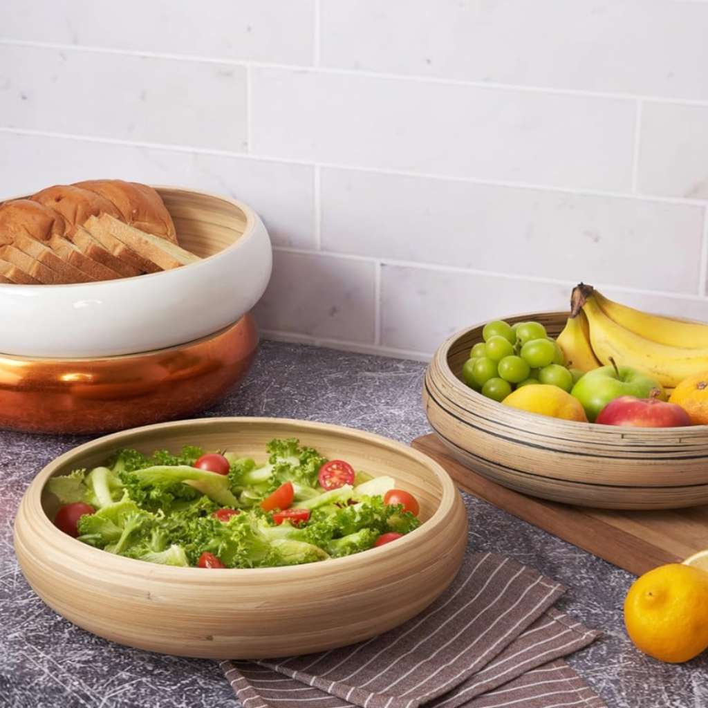 Natural bamboo salad bowl filled with fresh green salad including lettuce, tomatoes, and cucumbers, displayed on kitchen counter with fruits and bread in background